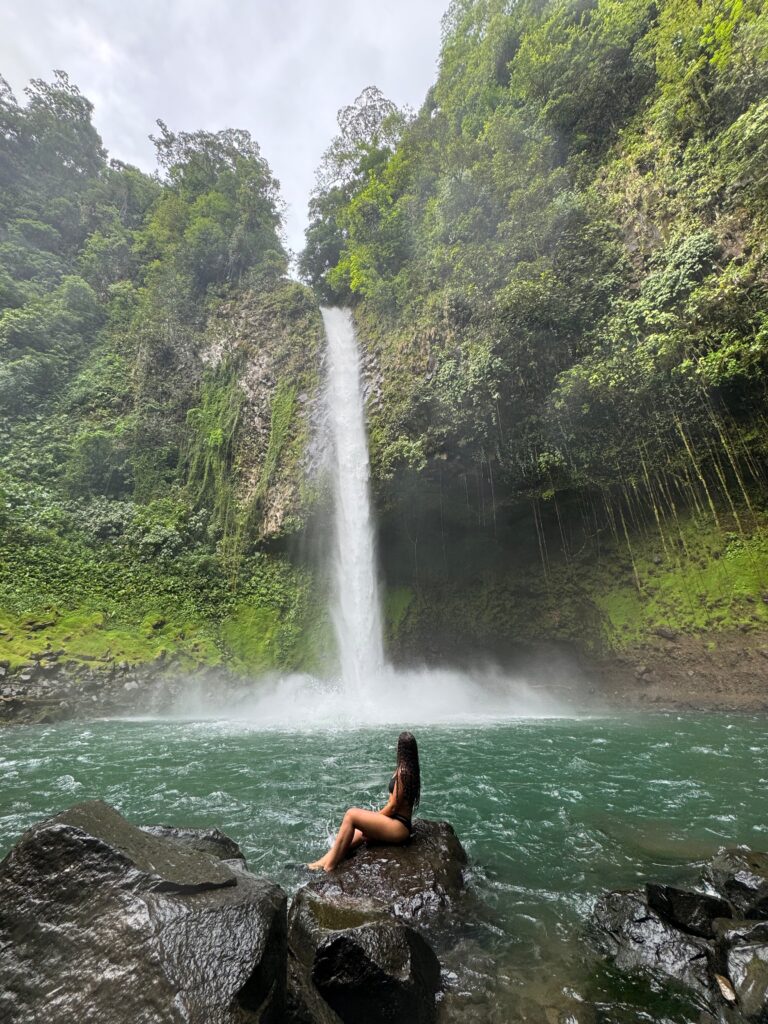 waterfall with blue water and rocks surrounding with girl in front. This is at La Fortuna waterfall in Costa Rica. One stop on 10 day itinerary.